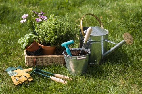A neatly maintained garden backyard features a lush, green lawn with a mix of grass textures, bordered by a flower bed containing potted plants with flowering and leafy greenery. In the foreground, gardening tools such as a pair of yellow gloves, a pair of pruning shears, and a small hand rake are placed on the grass. To the right, a metal watering can with a handle and spout is positioned next to a galvanized bucket, which holds a blue gardening trowel and a small spade. Behind the watering can, a rectangular wicker basket contains additional potted plants, some with purple and green foliage. The garden appears well-tended, with a clear separation between the flower bed and the lawn area, suggesting regular maintenance and plant care by a gardening service like Lawn Mowing Maida Vale. The scene is captured on a bright, partly cloudy day, with natural sunlight highlighting the vibrant green colours of the grass and plants, creating an inviting outdoor space suitable for gardening and lawn care activities in the Maida Vale area within postcode W9.